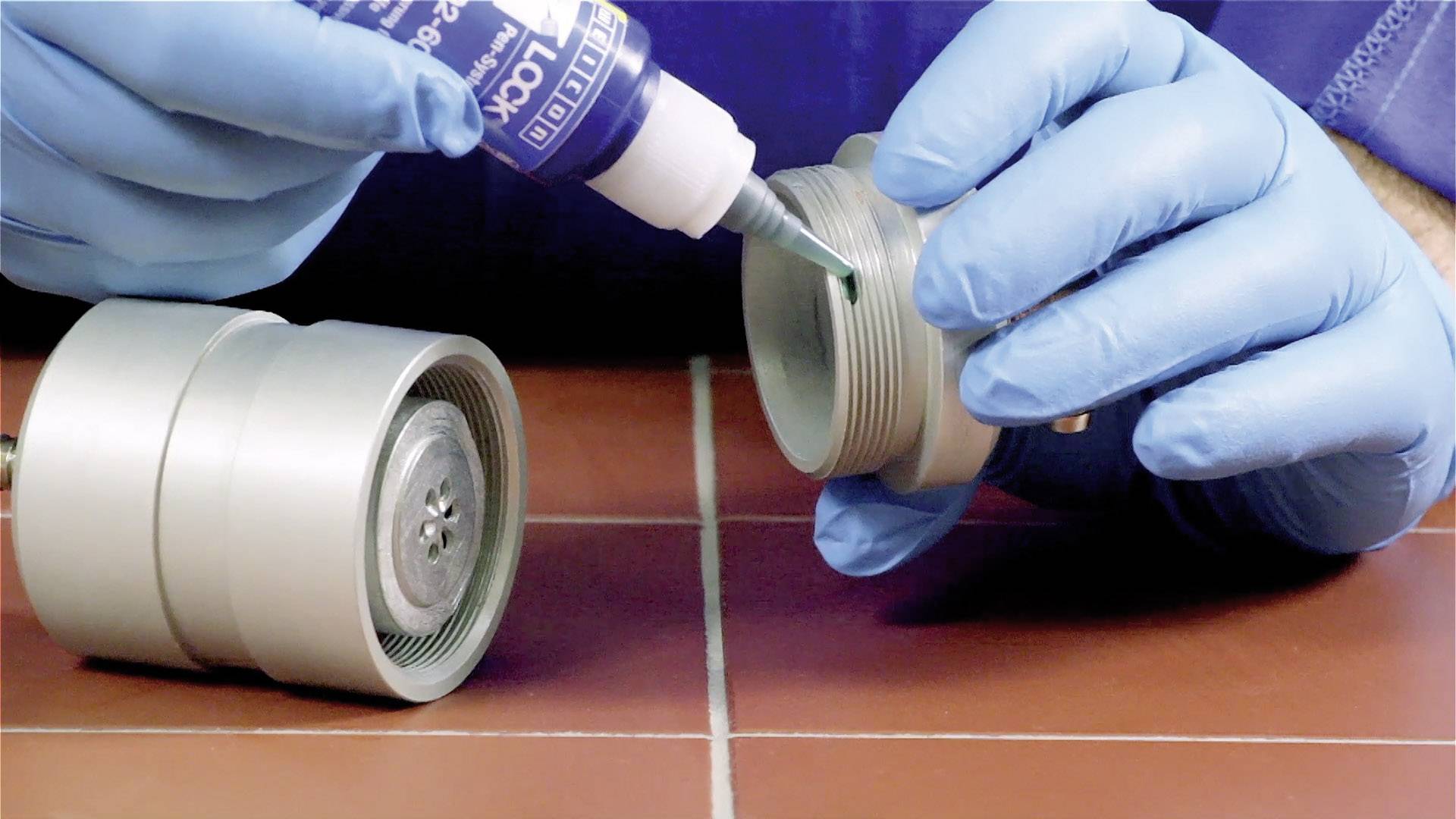 Una persona con guanti blu applica un sigillante liquido sui filetti di un cilindro metallico.