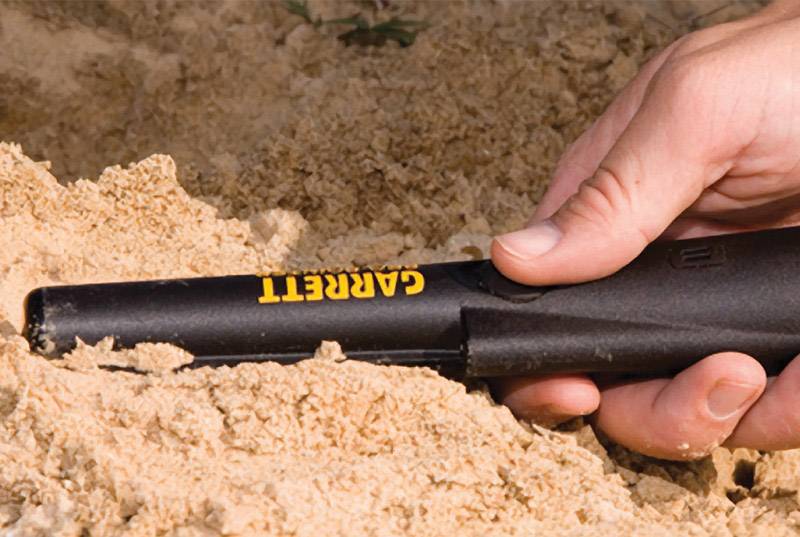 Una mano regge un metal detector, parzialmente sepolto nella sabbia. Lo strumento è nero con la scritta gialla 'Garrett'.