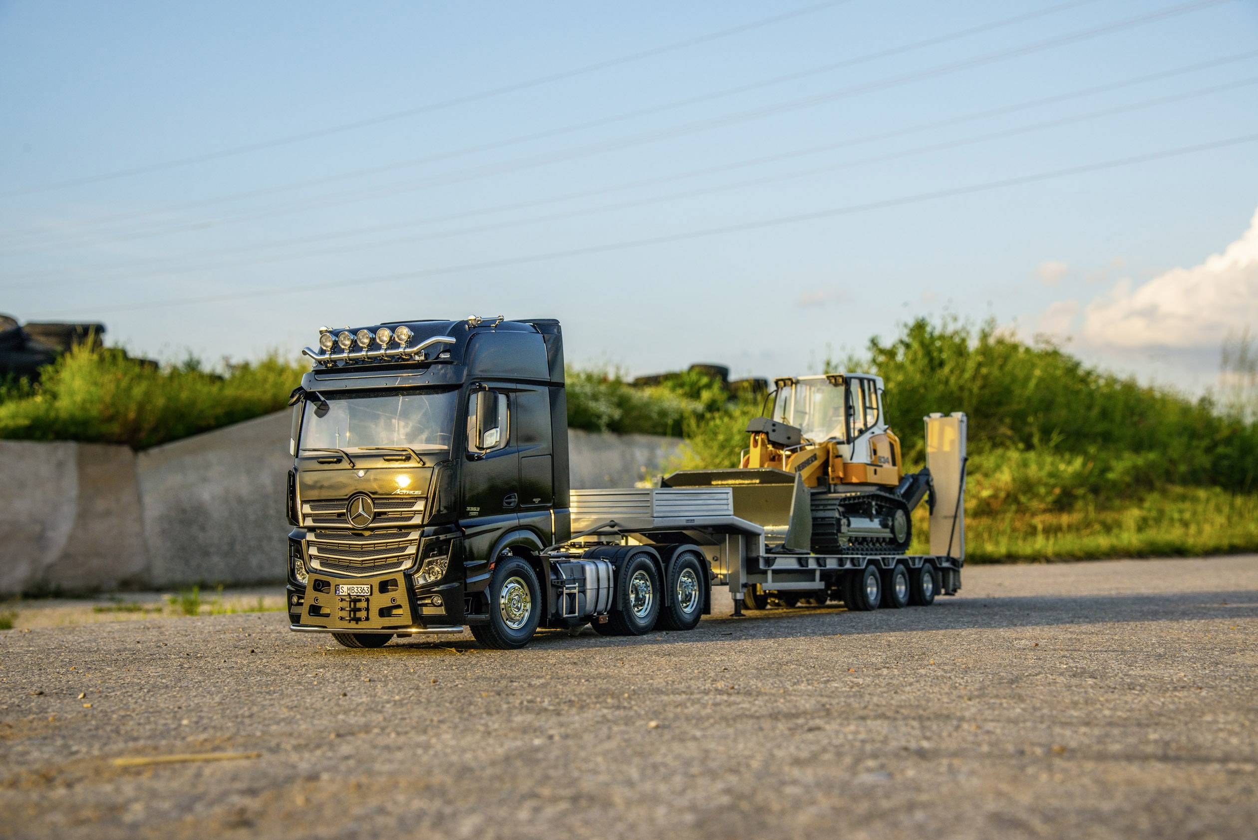 Un camion nero sta trasportando un rimorchio con un bulldozer giallo sopra, attraversando una superficie asfaltata, circondato da erba verde.