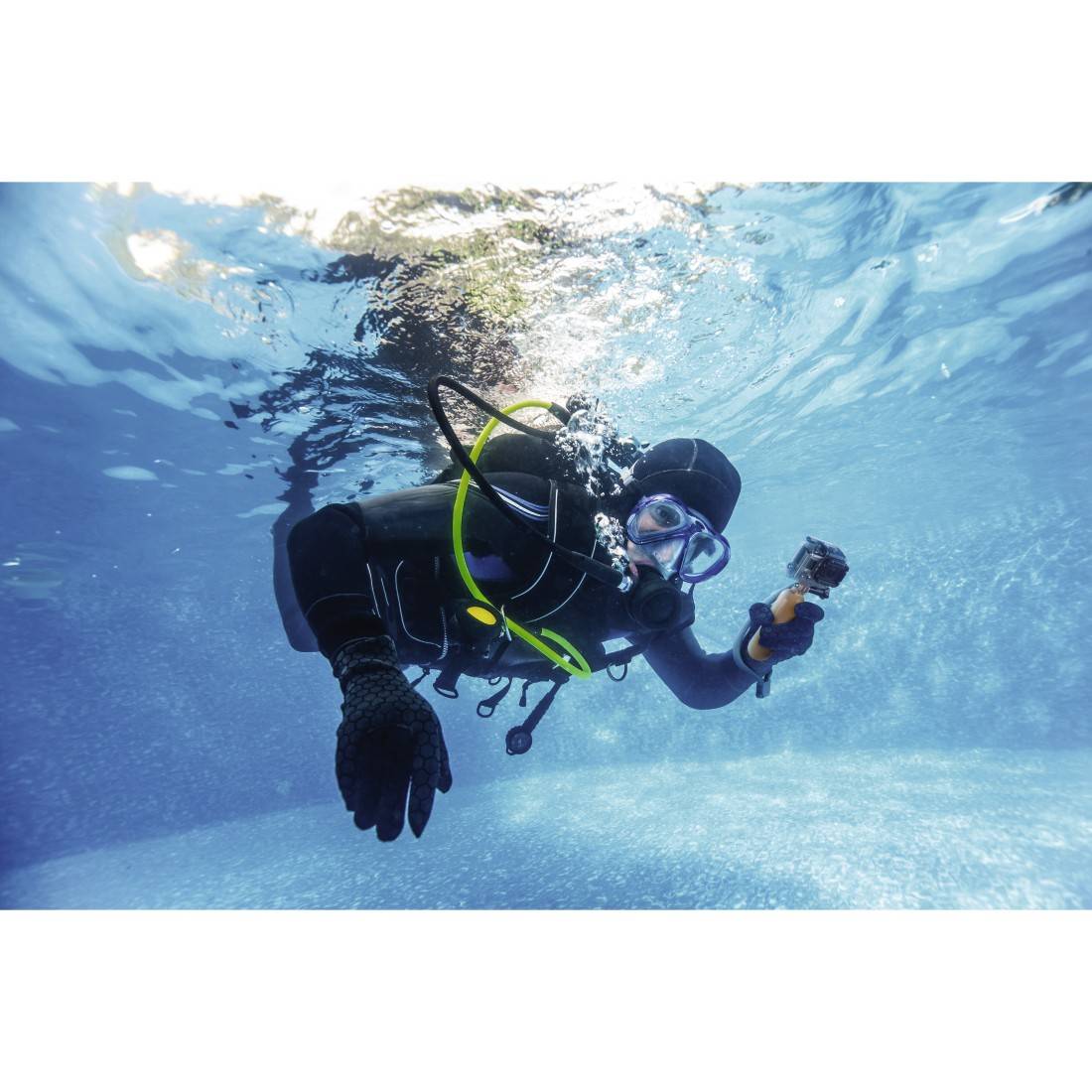 Un sommozzatore in muta da sub nuota sott'acqua tenendo in mano una macchina fotografica. Sullo sfondo un oceano blu cristallino.