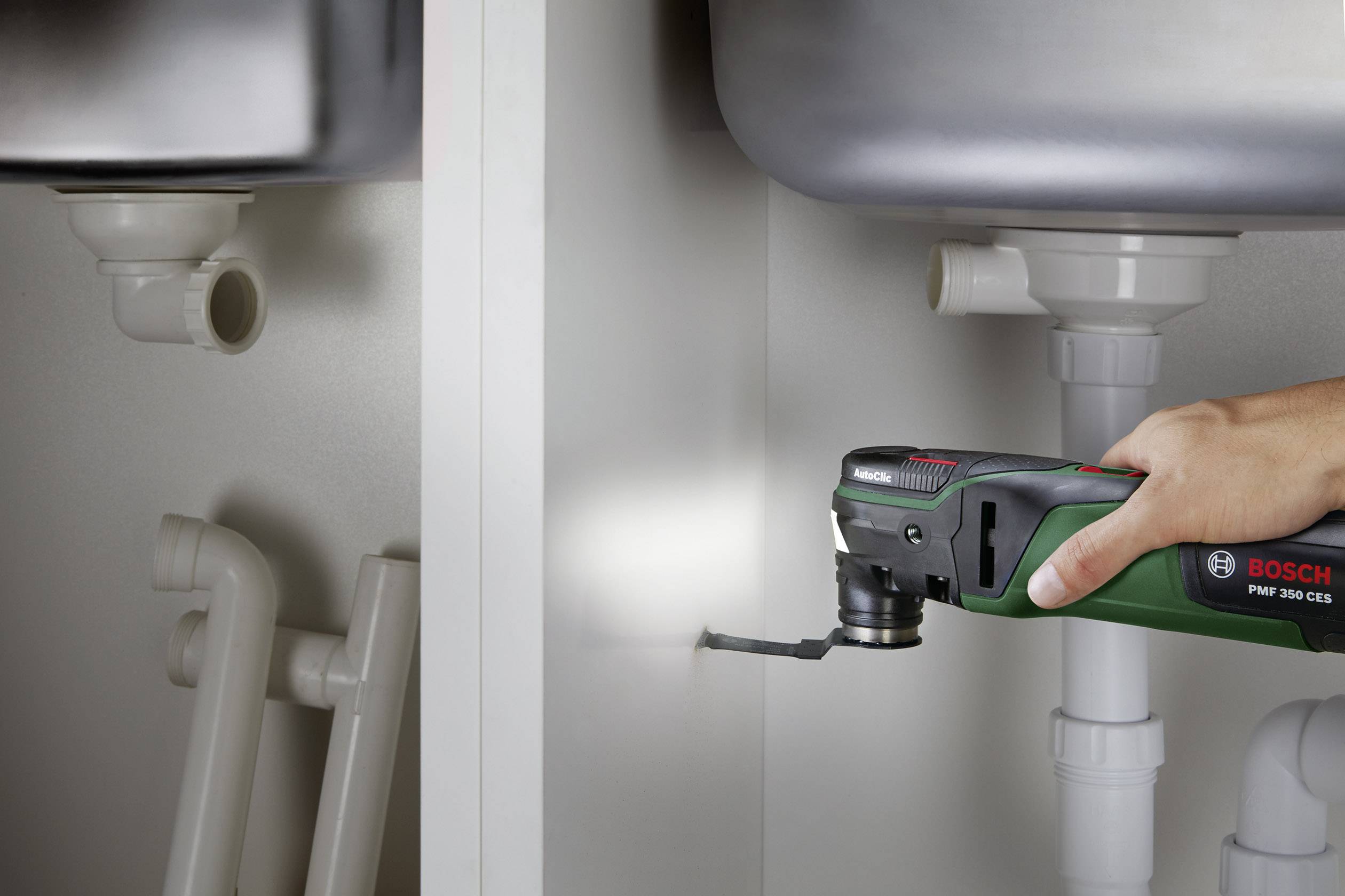 'Un utensile multiuso verde taglia un'asse di legno sotto un lavandino. Anche i tubi e il sifone sono visibili.'