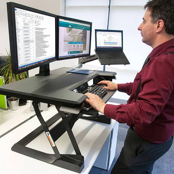 Una persona lavora in ufficio a una scrivania regolabile in altezza con due monitor e un laptop, entrambi gli schermi mostrano pagine web.