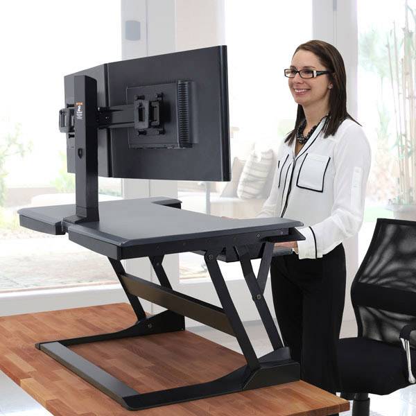 Una donna sorride accanto a una scrivania regolabile in altezza davanti a uno schermo del computer in un ufficio moderno.