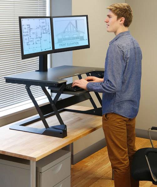 Una persona è in piedi a un tavolo regolabile in altezza e lavora su due monitor che mostrano progetti architettonici.