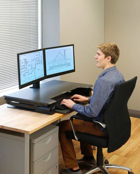 Un uomo è seduto a una scrivania e sta lavorando su un computer con due monitor. Sugli schermi sono visibili dei progetti di costruzione.