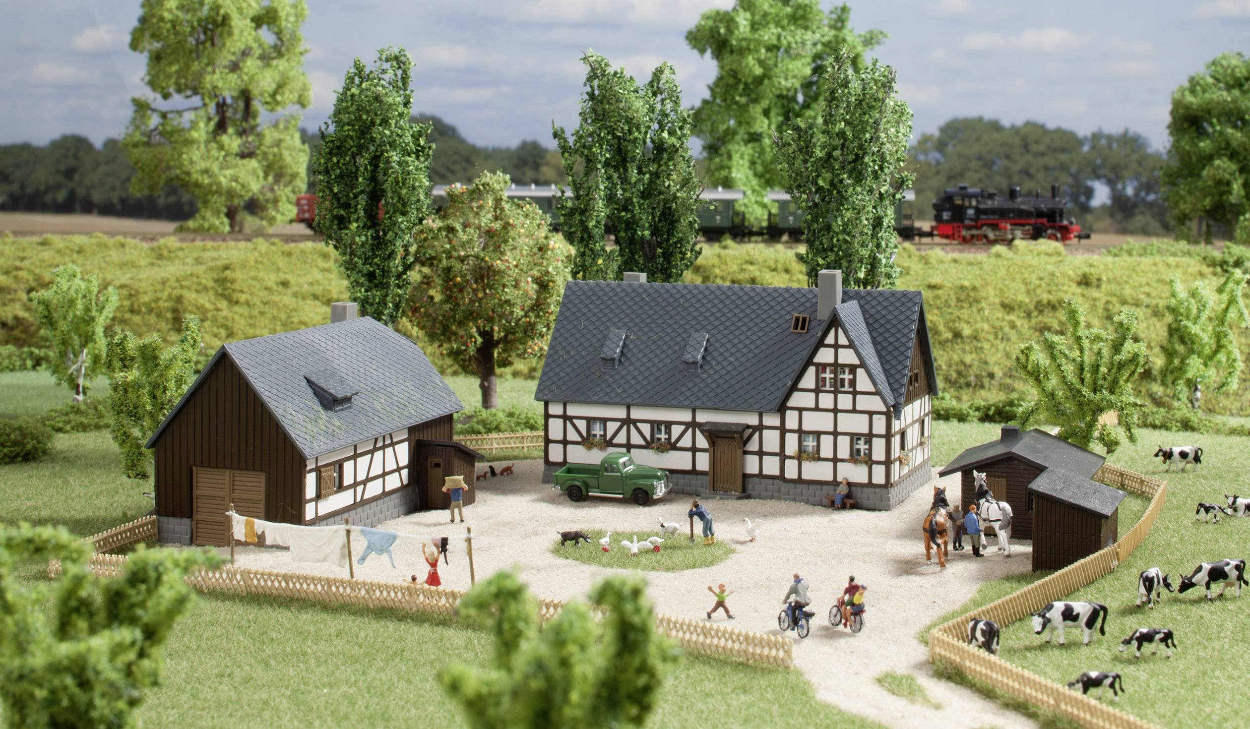 Un paesaggio in miniatura mostra un'azienda agricola con due case a graticcio, circondata da prati e alberi. Le mucche pascolano nell'area recintata. Sullo sfondo, un treno passa.