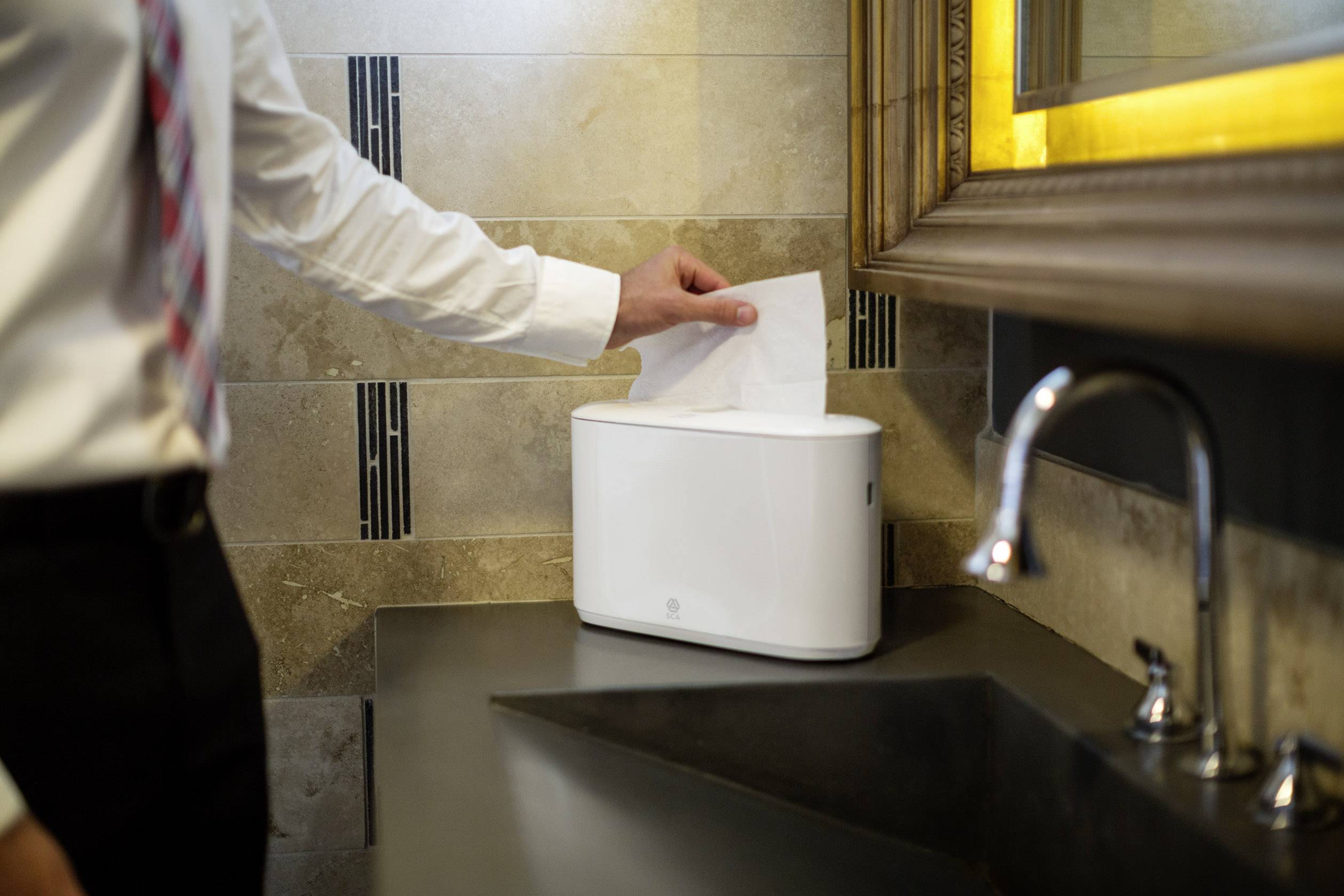 Un uomo in giacca e cravatta inserisce un asciugamano di carta in un dispenser moderno per asciugamani di carta in un bagno arredato con stile.