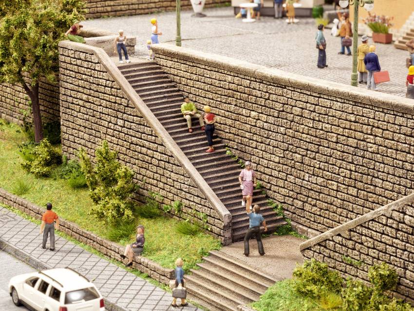 Una scena in miniatura dettagliata mostra una scala in pietra su un muro con delle persone, alcune sedute, altre che salgono o scendono le scale. In primo piano c'è un'auto bianca sulla strada.