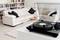 Giradischi con disco in vinile nero sullo sfondo di un soggiorno moderno con divani beige e libreria sullo sfondo.