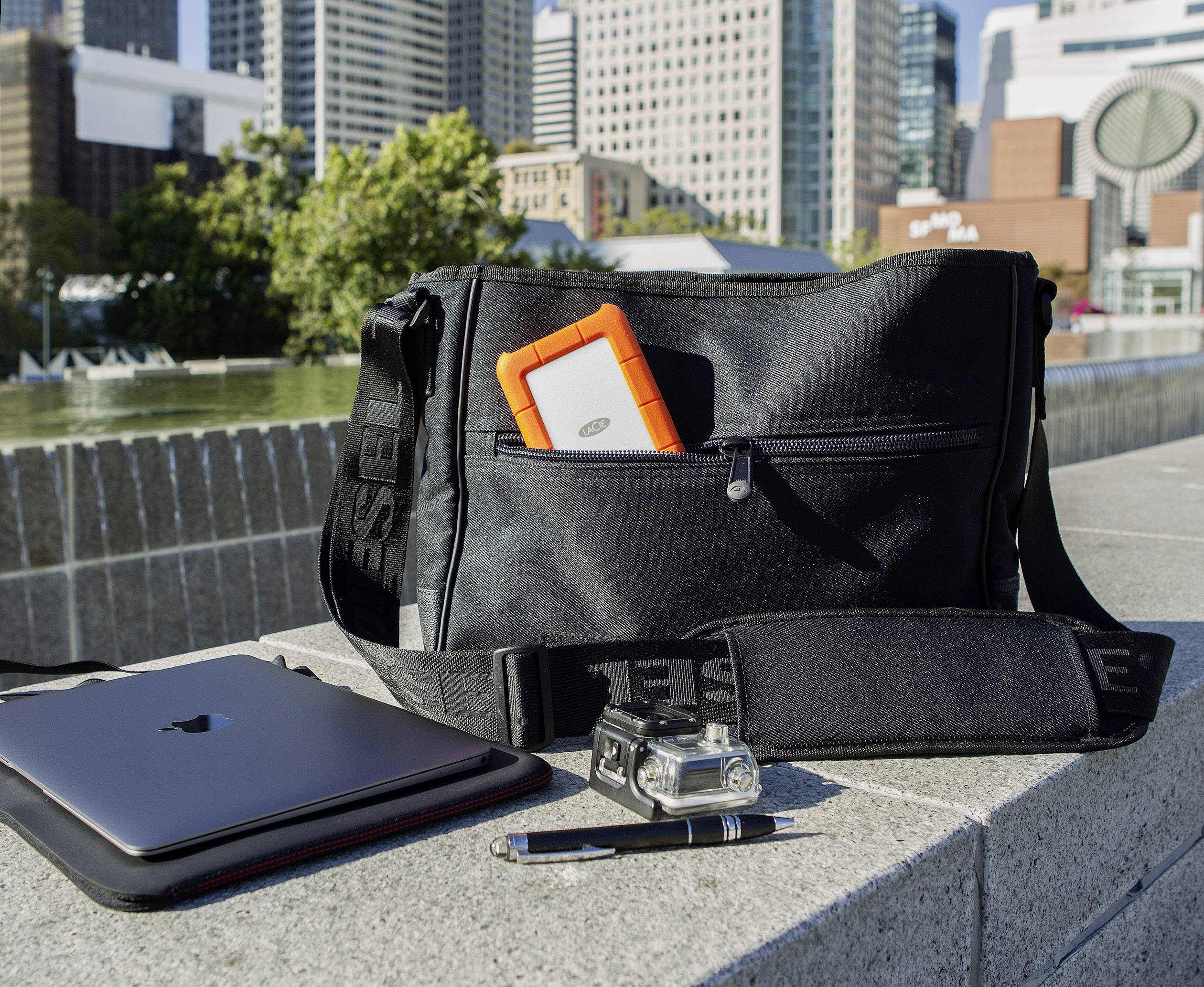 Una borsa nera con un hard disk arancione, un laptop, una fotocamera e una penna in un ambiente urbano all'aperto.