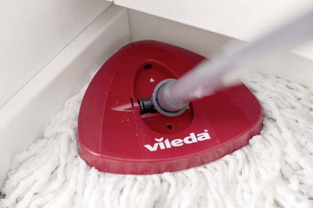 Una testina rossa di un mop con il marchio 'Vileda' in un angolo su un pavimento bianco.