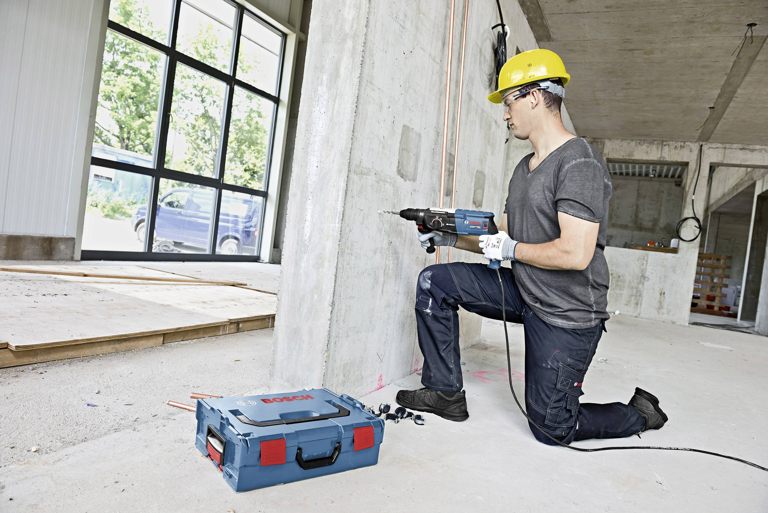 Un operaio edile è inginocchiato sul pavimento di un edificio in costruzione e sta perforando dei fori in un muro di cemento con un utensile elettrico.