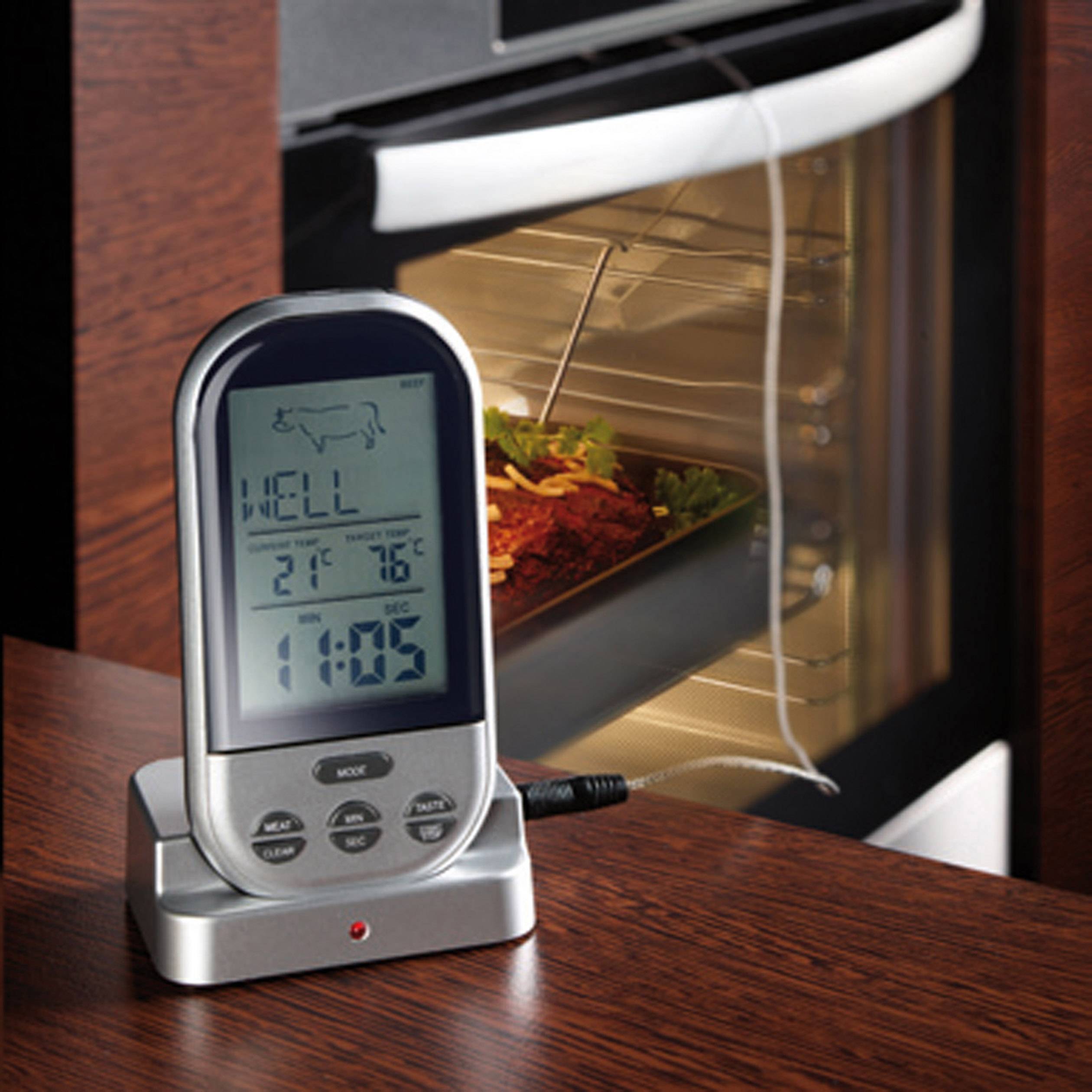 Un termometro digitale per la carne su un tavolo mostra 'WELL' e temperature accanto a un forno pieno. Un cavo conduce all'interno del forno.