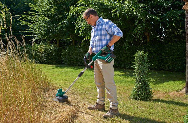 Un uomo taglia l'erba alta con un decespugliatore elettrico in giardino. Indossa pantaloni casual e una camicia a quadri.