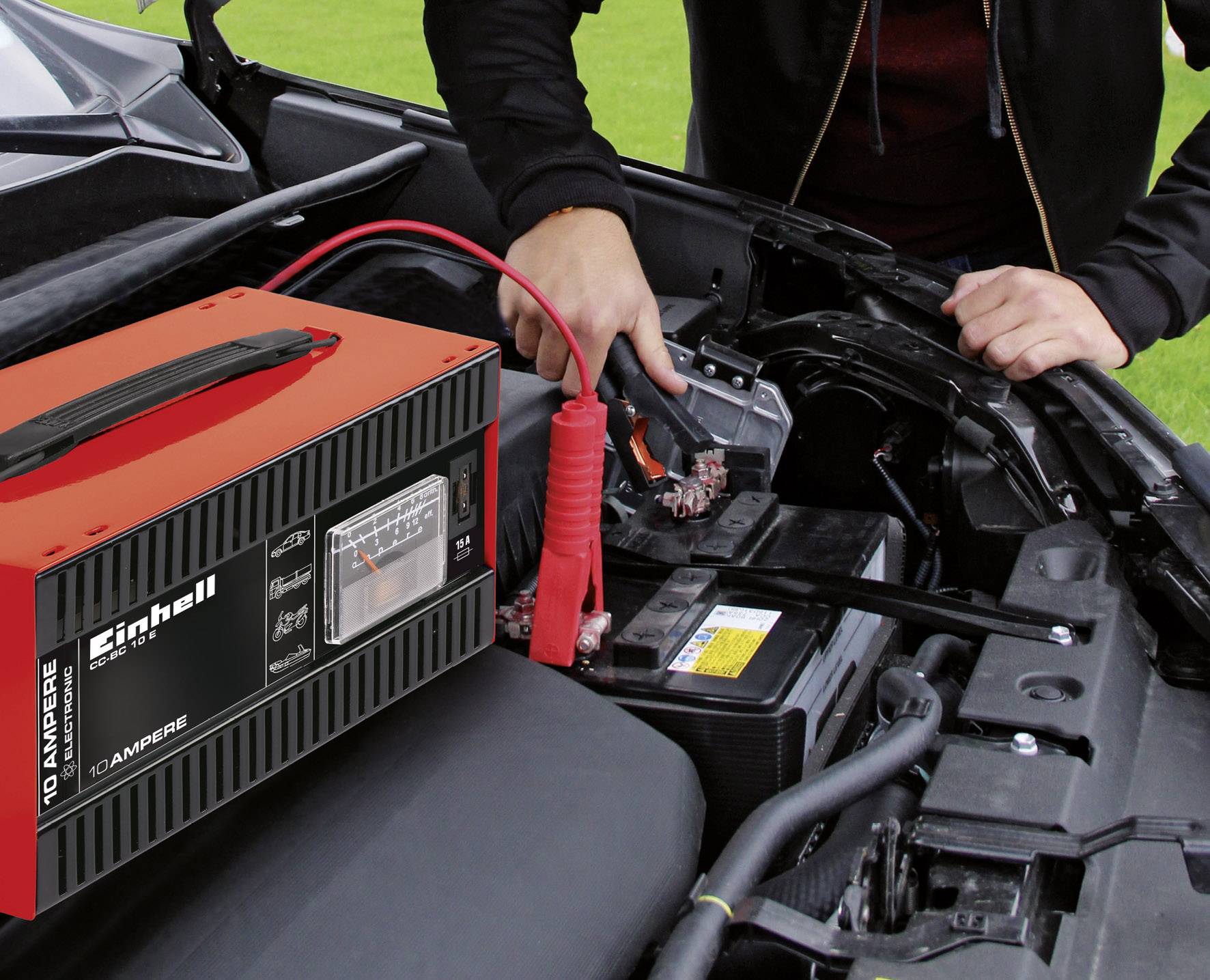 Persona che fa il ponte della batteria dell'auto utilizzando cavi per l'avviamento, con un caricabatterie rosso in primo piano. Sullo sfondo un prato con un cofano motore aperto.