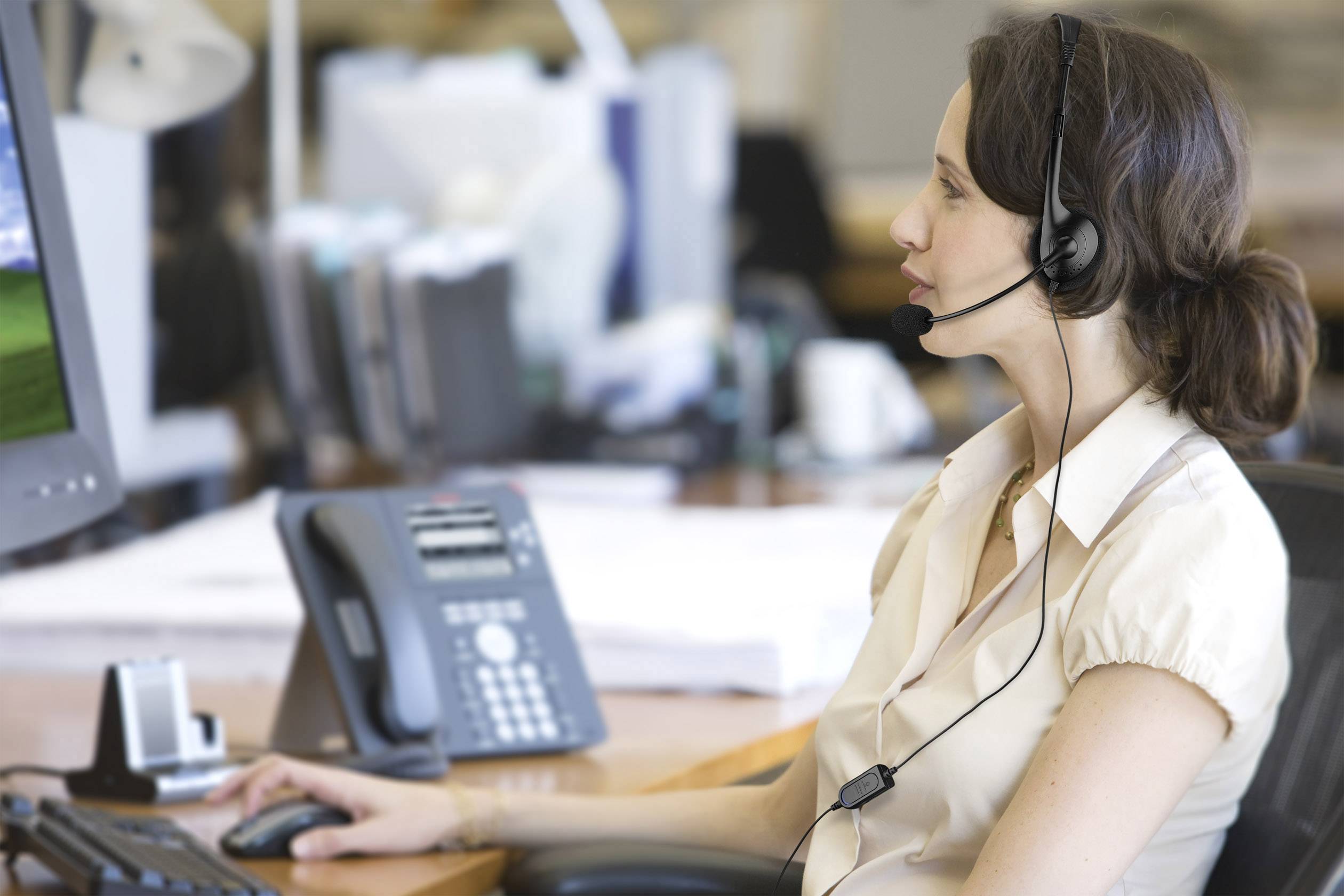 Una donna con un headset lavora concentrata al computer in un ufficio. Sullo sfondo si vedono un telefono da tavolo e altri elementi di ufficio sfocati.