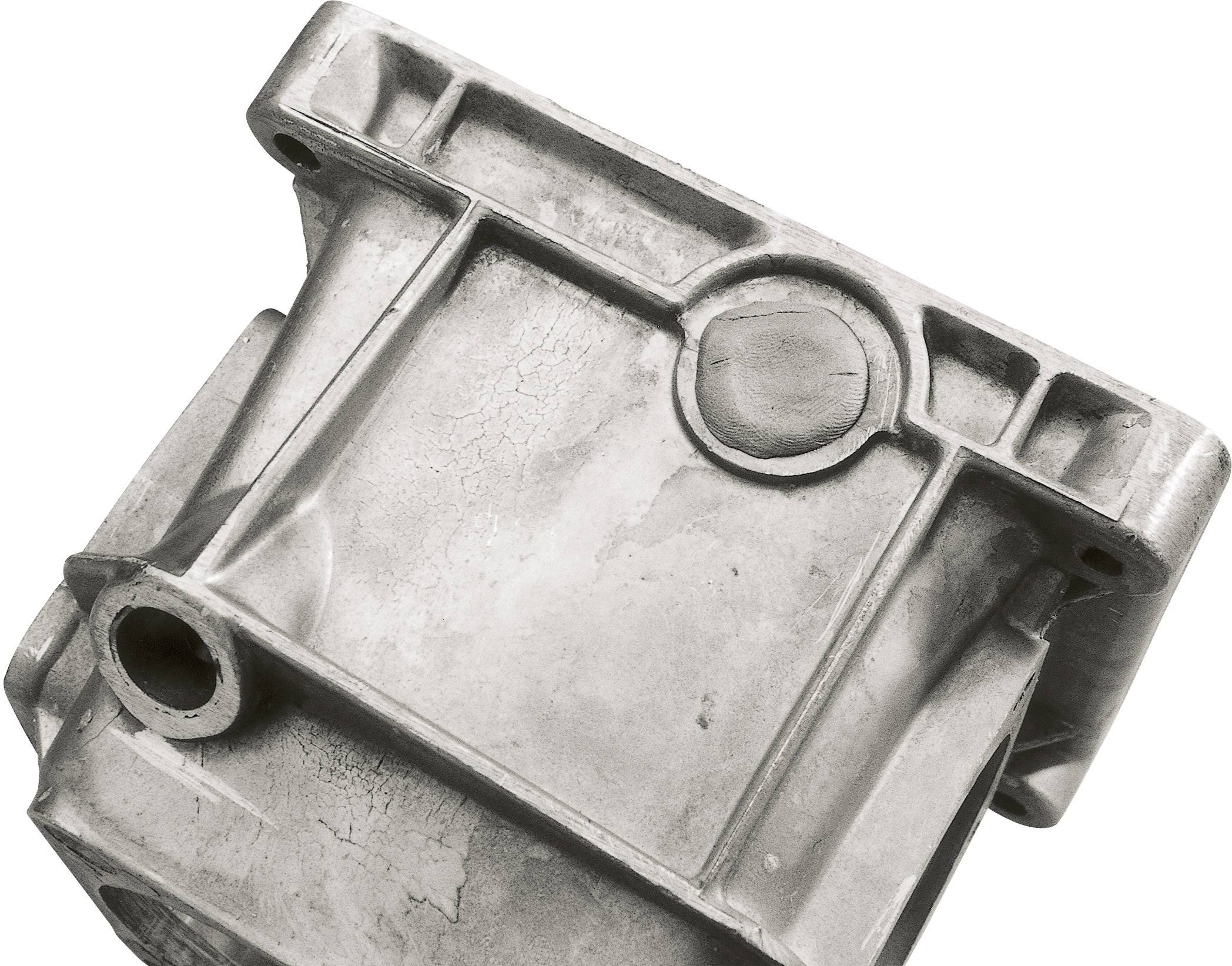Un componente rettangolare in metallo con diverse camere e un rilievo circolare. Appare industriale e mostra segni di usura.