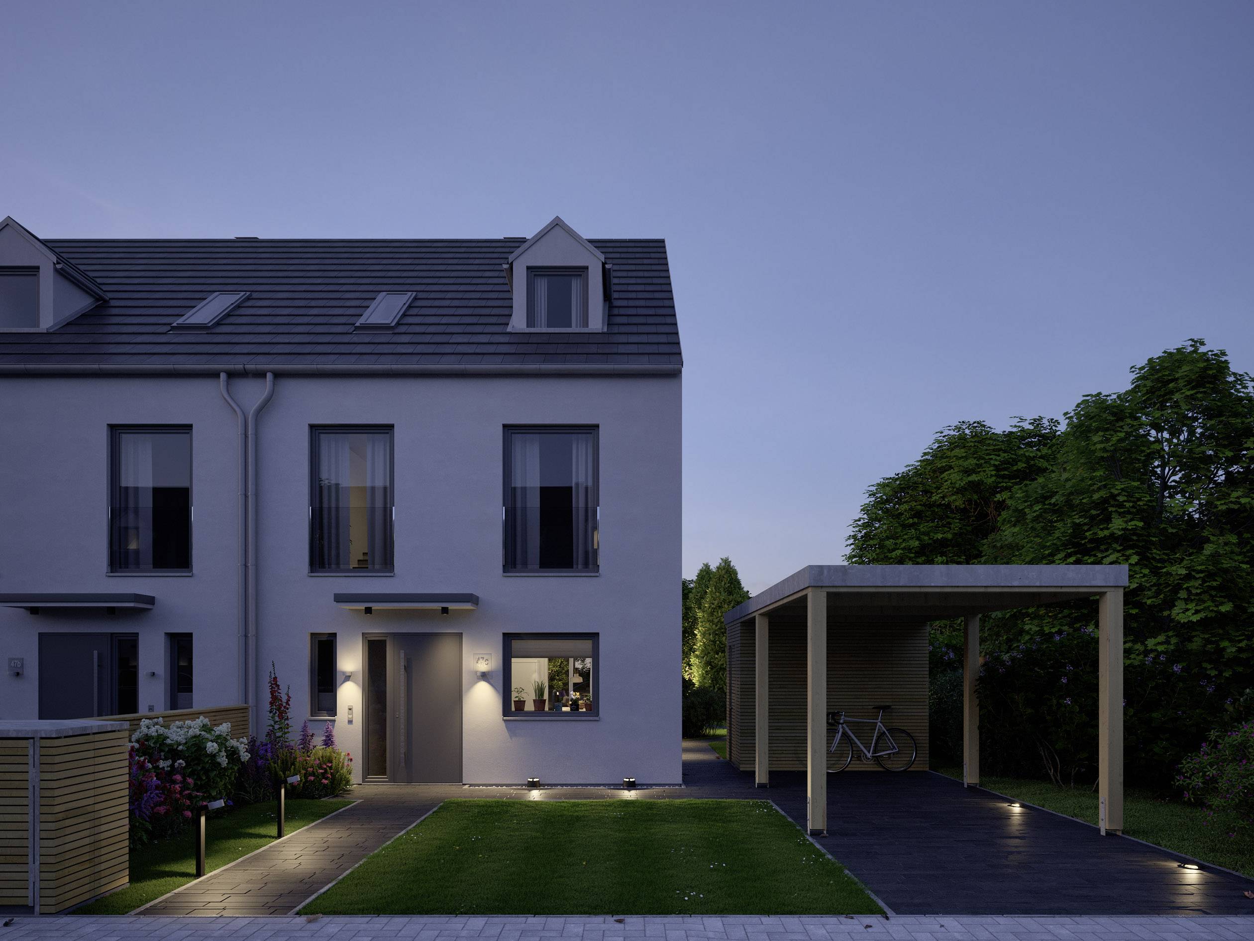 Casa bifamiliare su due piani al crepuscolo con ingresso illuminato, giardino curato e un carport coperto sulla destra dell'immagine.