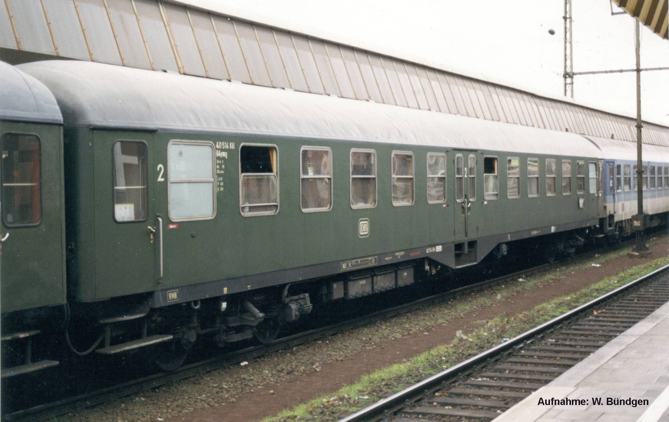 Un vagone ferroviario verde è fermo su un binario. Ha diversi finestrini e la scritta '2a classe'. Sullo sfondo si vede un edificio della stazione ferroviaria.