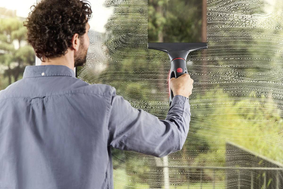 Un uomo sta pulendo una finestra con un tergivetro. Fuori si vedono alberi e un giardino.