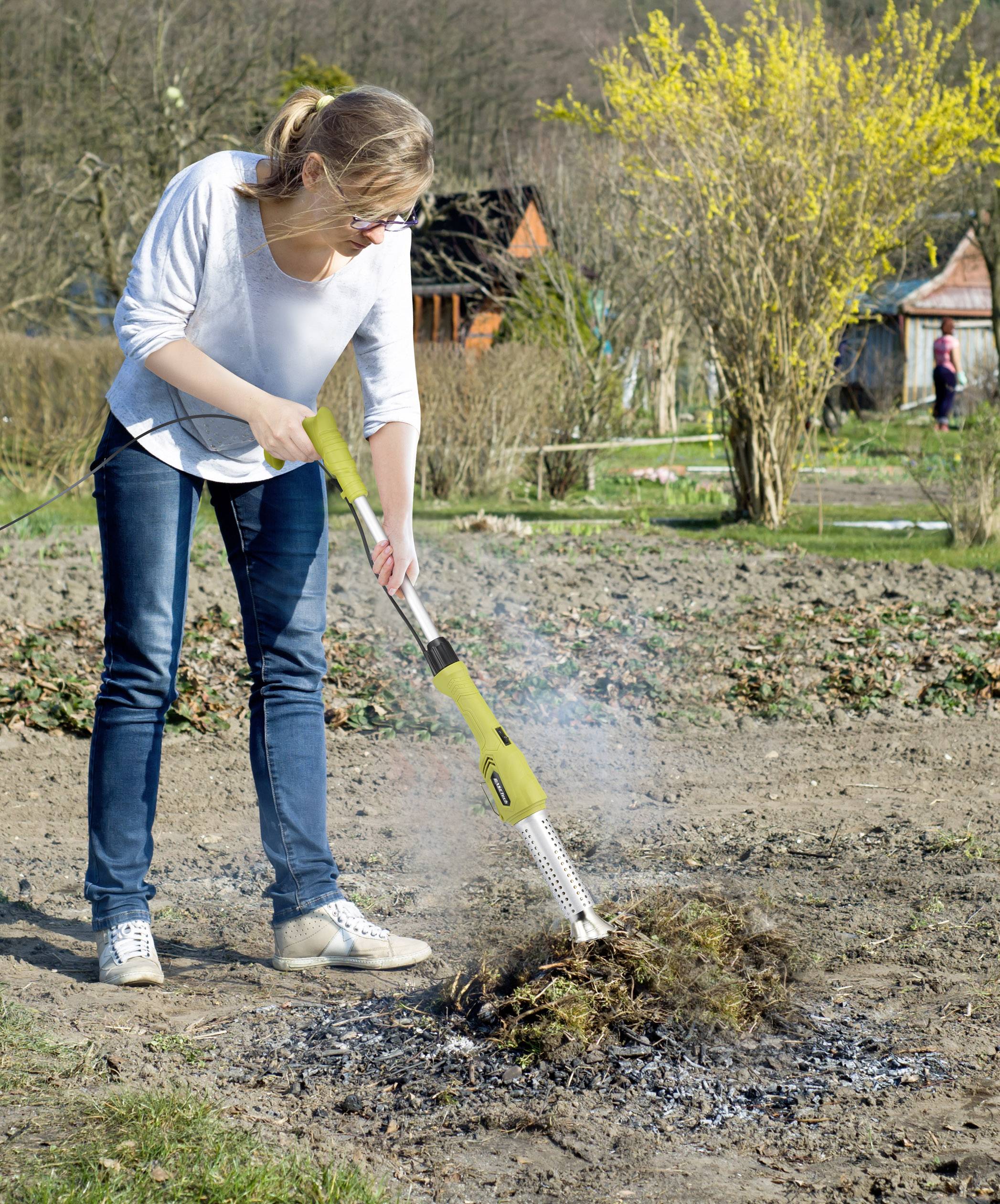 Una donna sta usando un attrezzo da giardino per bruciare le erbacce in un giardino. Sullo sfondo si vedono alberi e cespugli.