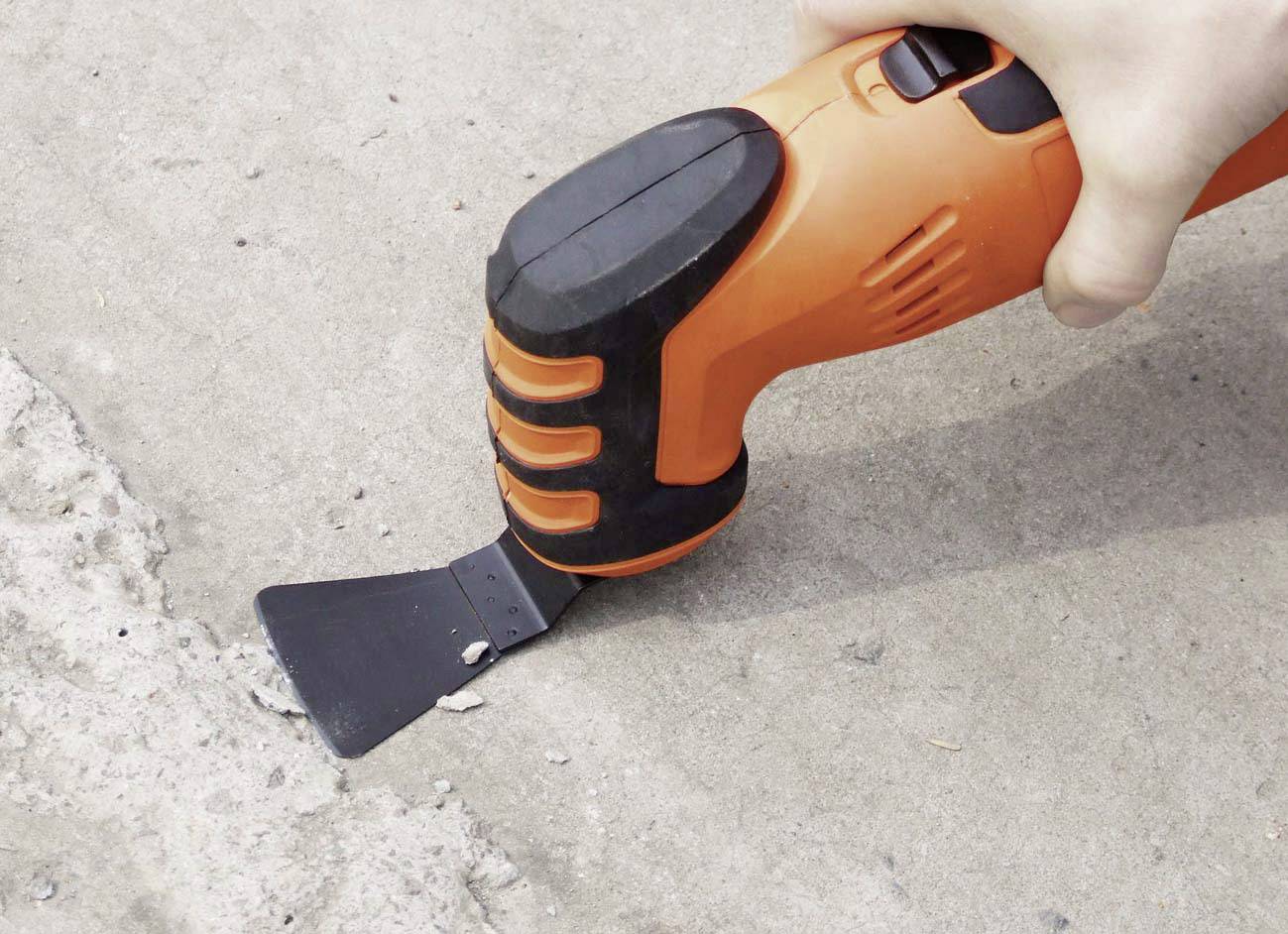 Una mano tiene un utensile elettrico arancione con un accessorio per spatola che rimuove il calcestruzzo da una superficie.
