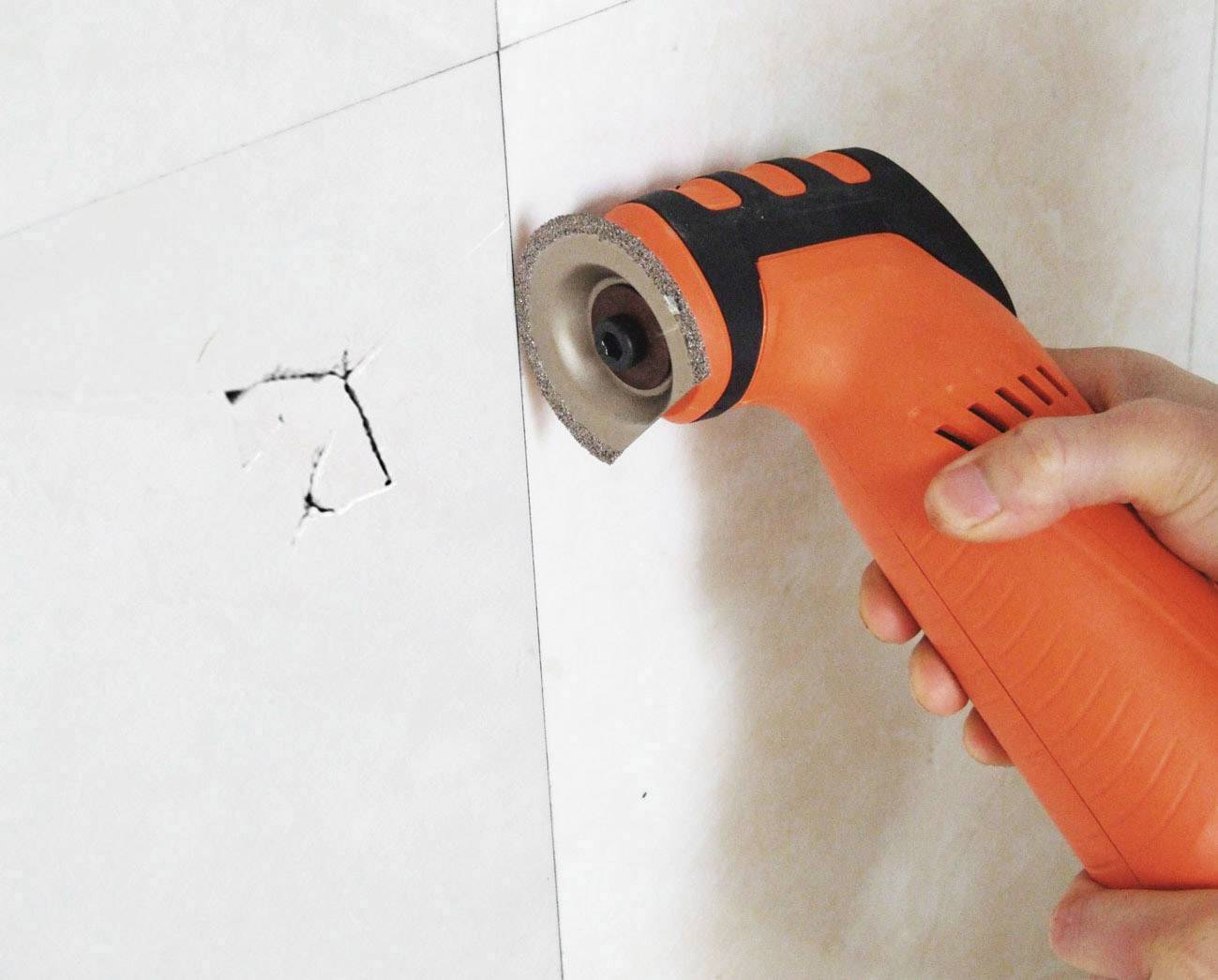 Una mano impugna un utensile elettrico arancione e taglia un piccolo foro in un muro o una piastrella bianca.