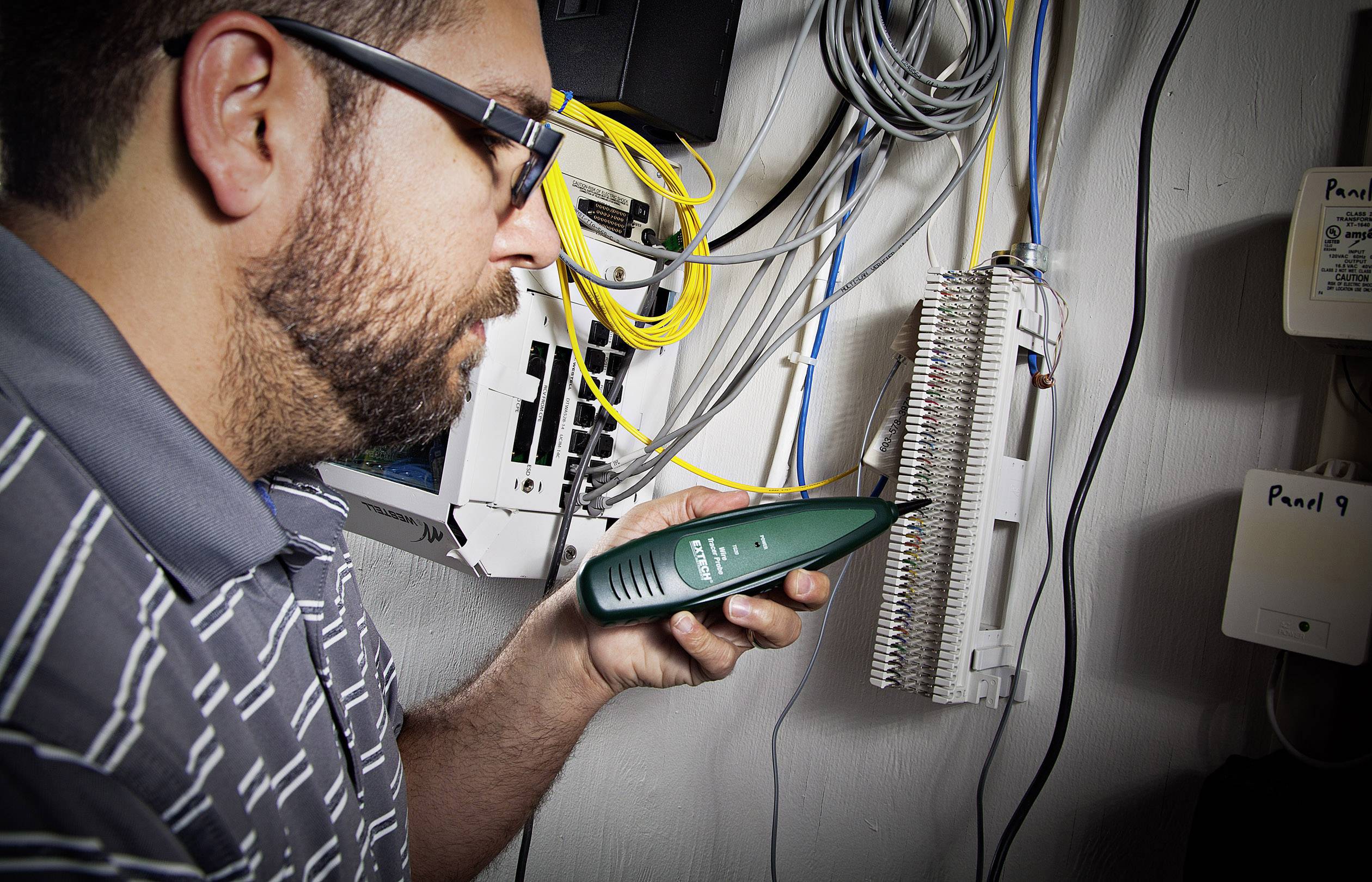 Un uomo sta controllando i cablaggi in un quadro elettrico con un dispositivo di verifica verde, circondato da diversi cavi.