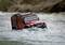 Un'auto giocattolo rossa di un fuoristrada guida attraverso acqua bassa, circondato da un fiume che scorre.