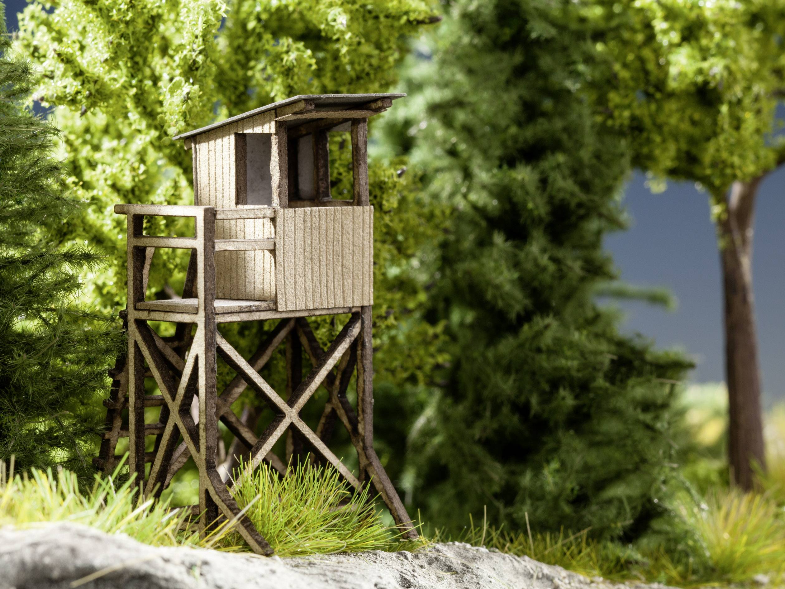 Una torre di legno per la caccia si erge nel bosco, circondada da alberi e cespugli verdi, sullo sfondo di un cielo blu.