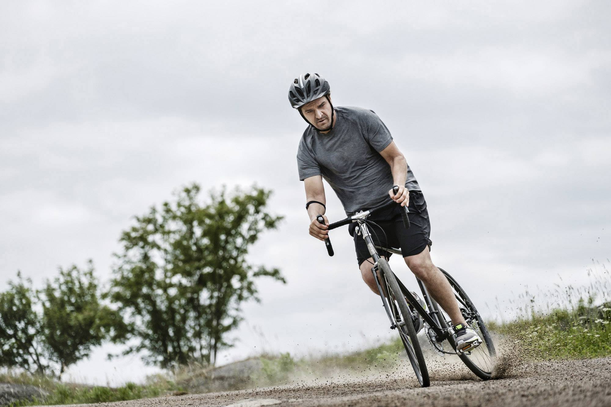 Una persona sta pedalando con una bicicletta lungo un sentiero di campagna. Sullo sfondo si vedono alberi e cielo.