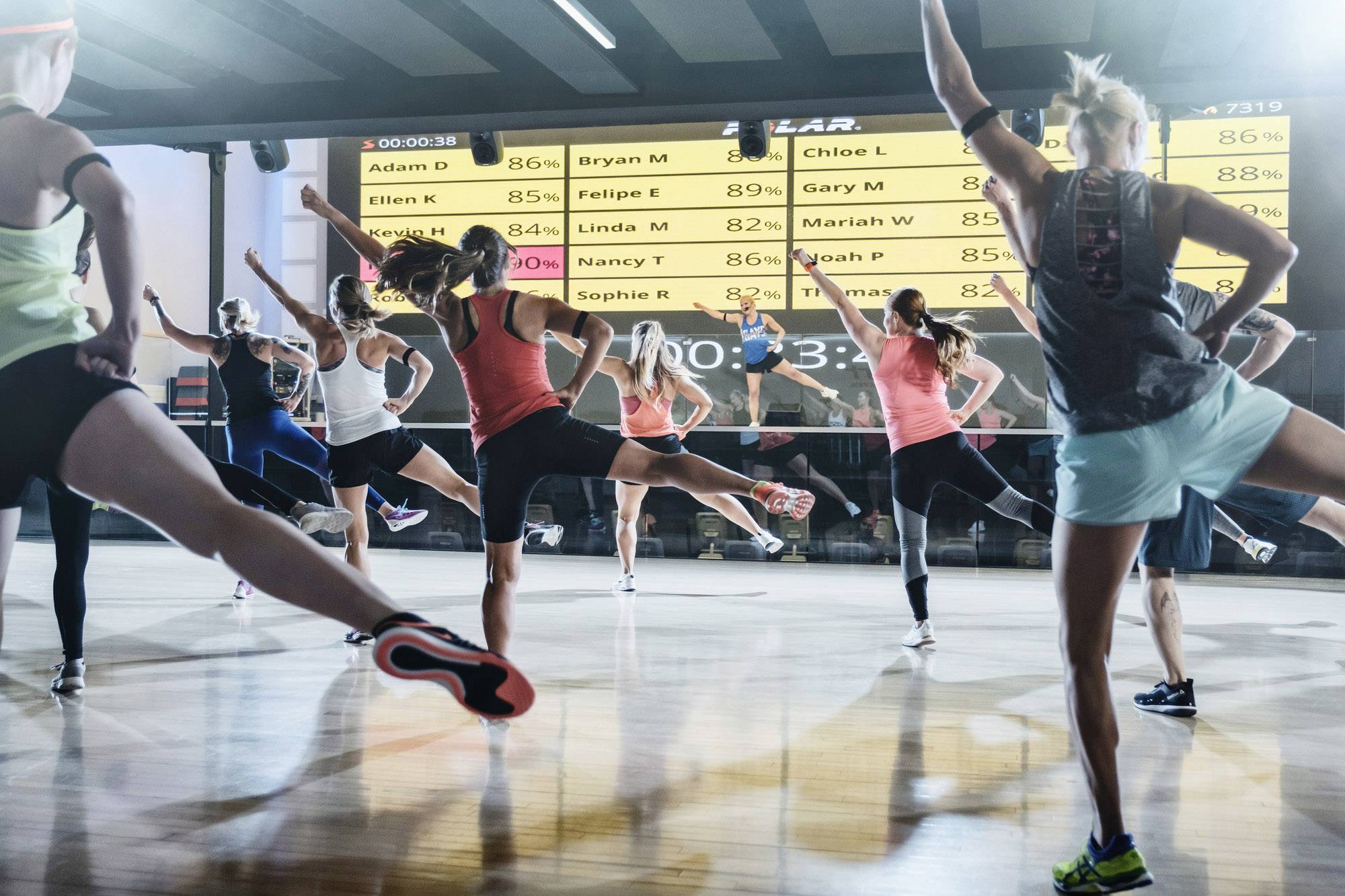 Un gruppo di persone partecipa a un corso di aerobica in una palestra. Sullo sfondo, uno schermo mostra i risultati delle prestazioni.