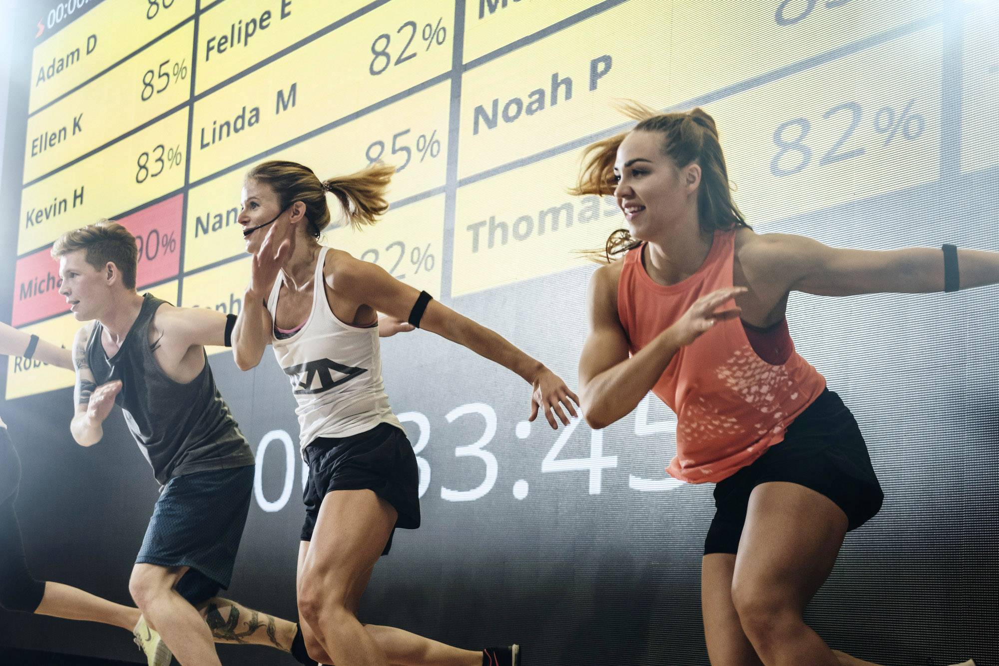 Diverse persone si allenano in una palestra. Sullo sfondo, si vedono schermi con nomi e percentuali.