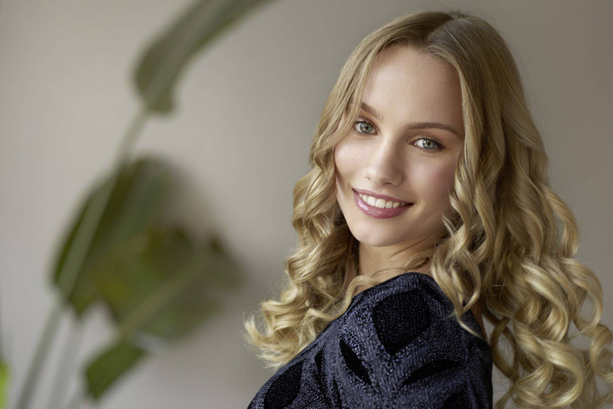 Una persona sorridente con capelli ricci indossa una maglia nera. Sullo sfondo si vedono foglie verdi sfocate.