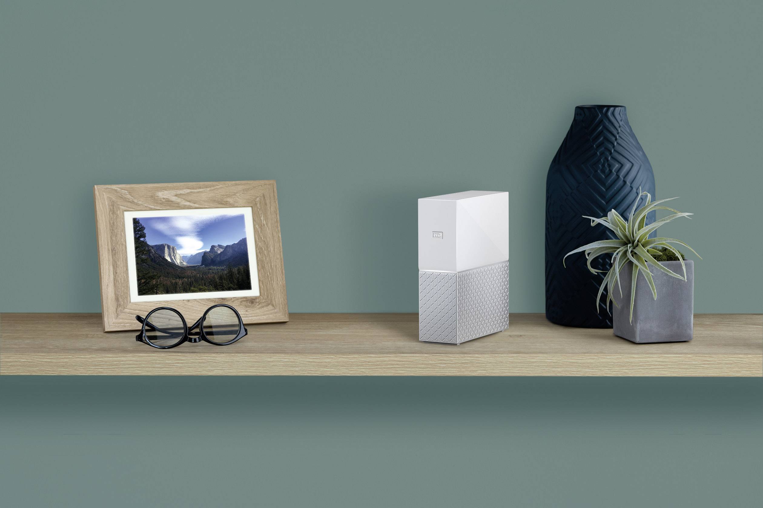 Una mensola con occhiali, cornice per foto con paesaggio, dispositivo di archiviazione digitale e vaso di piante sullo sfondo verde.