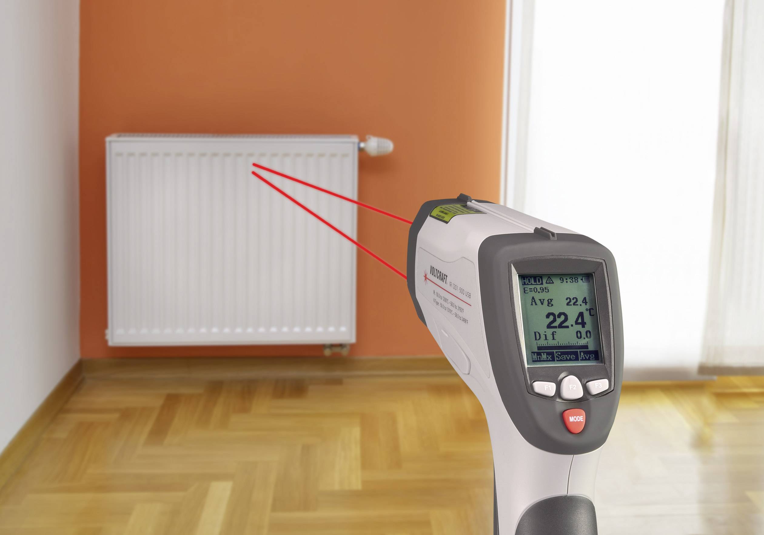 Un termometro a infrarossi misura la temperatura superficiale di un radiatore in una stanza con pavimento in legno e parete arancione.