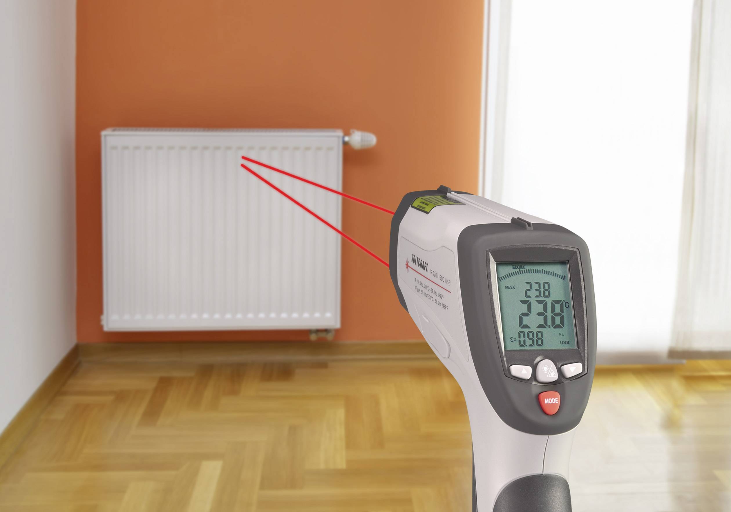 Un termometro a infrarossi con raggi laser misura la temperatura di un radiatore in una stanza con pavimento in legno e parete arancione.