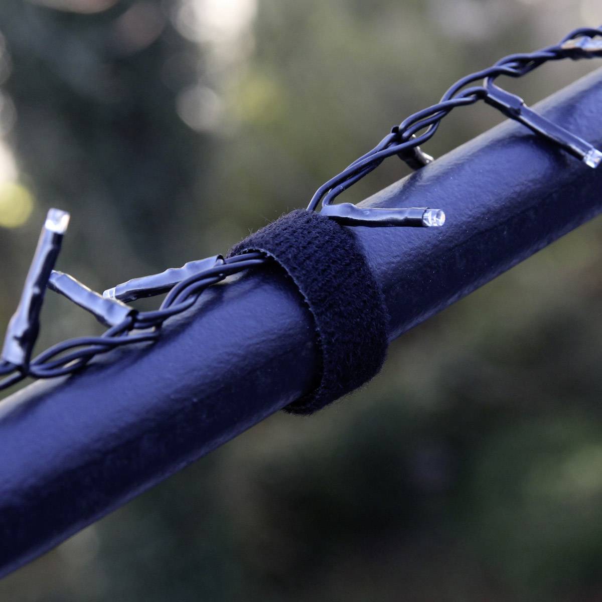 Catena luminosa fissata con un nastro di velcro nero su un corrimano scuro, con piante sfocate sullo sfondo.