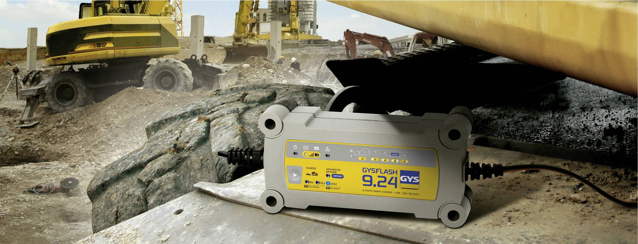 Un caricabatterie GYSFLASH 9.24 è posizionato su un pezzo di metallo in un cantiere edile. Sullo sfondo si vedono attrezzature da costruzione e movimenti di terra.