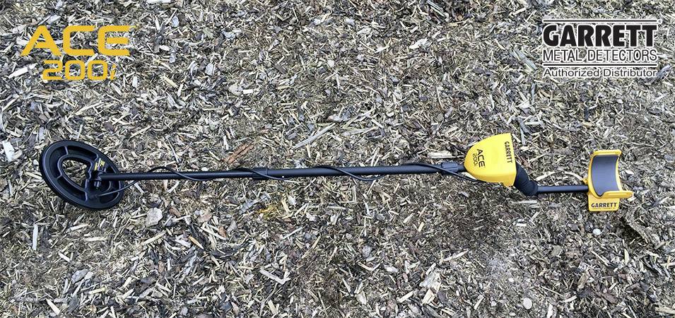 Metaldetector ACE 200 poggia sul terreno boschivo. Il dispositivo è nero e giallo con la scritta 'Garrett Metal Detectors' in alto a destra nell'immagine.
