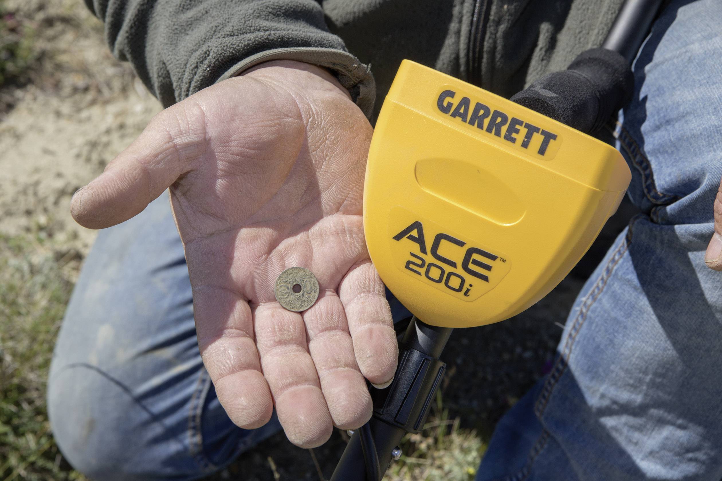Una persona tiene in mano una piccola moneta antica e nell'altra mano tiene un metal detector. Il detector è giallo e reca la scritta 'ACE 200i'.