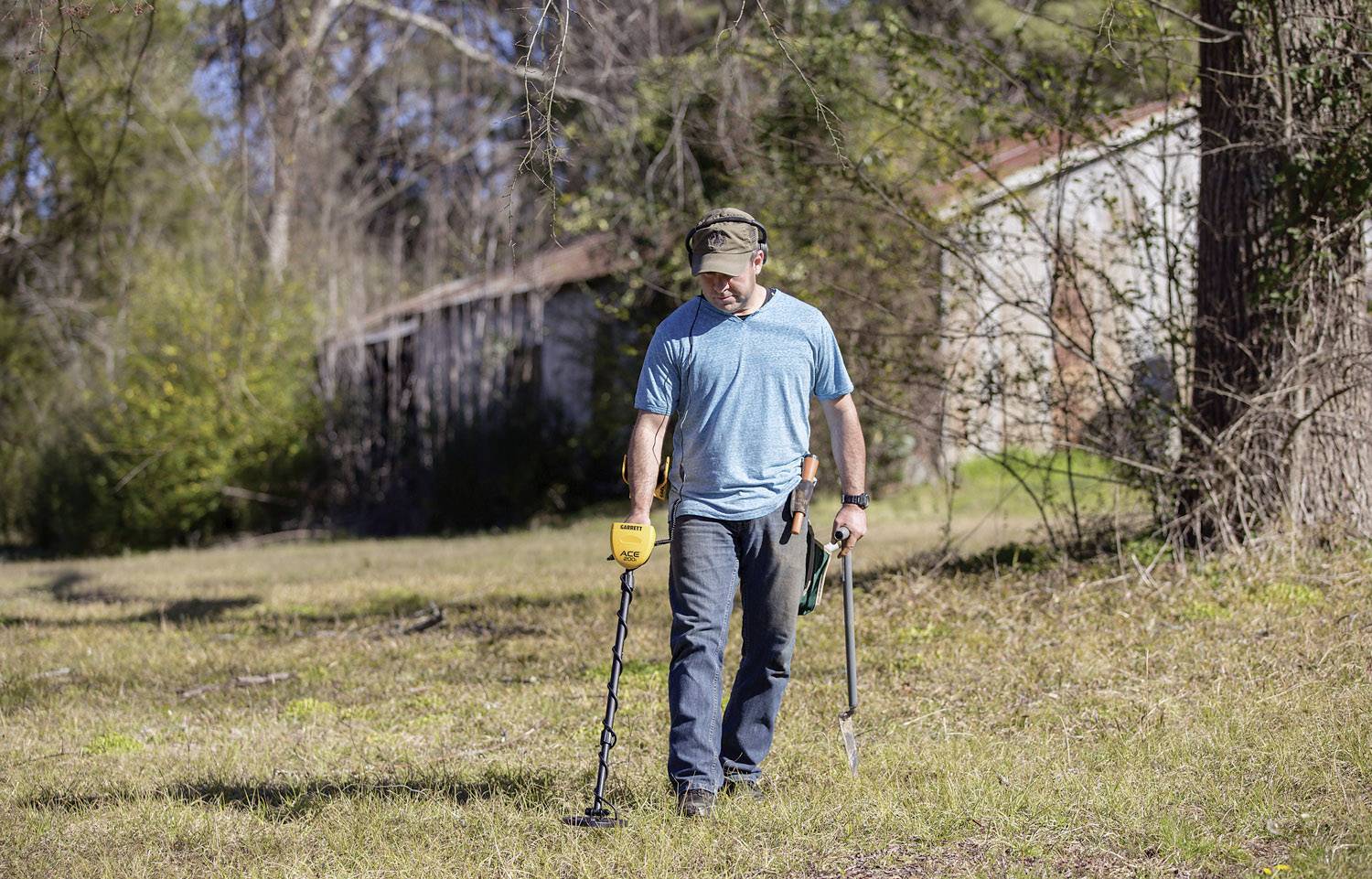 Persona con metal detector che cerca su un campo erboso. Tiene una pala in mano. Sullo sfondo si vedono alberi e un edificio.