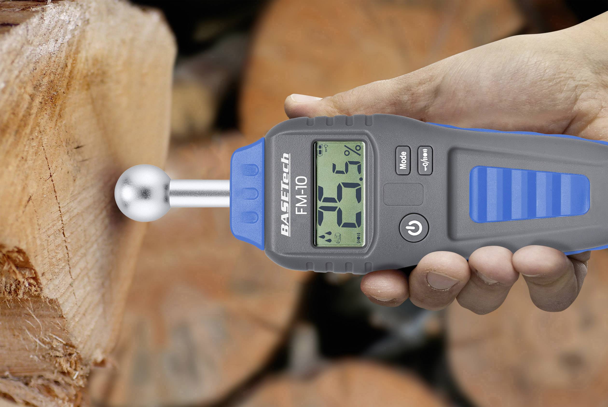 Un misuratore di umidità mostra '20%' mentre è appoggiato su un pezzo di legno. Sullo sfondo si vedono dei tronchi di legno.