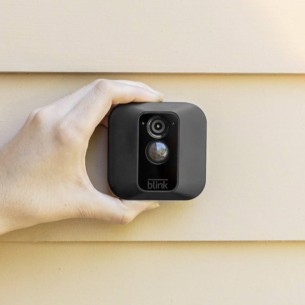 Una mano tiene e installa una piccola telecamera di sicurezza nera su una parete esterna chiara.