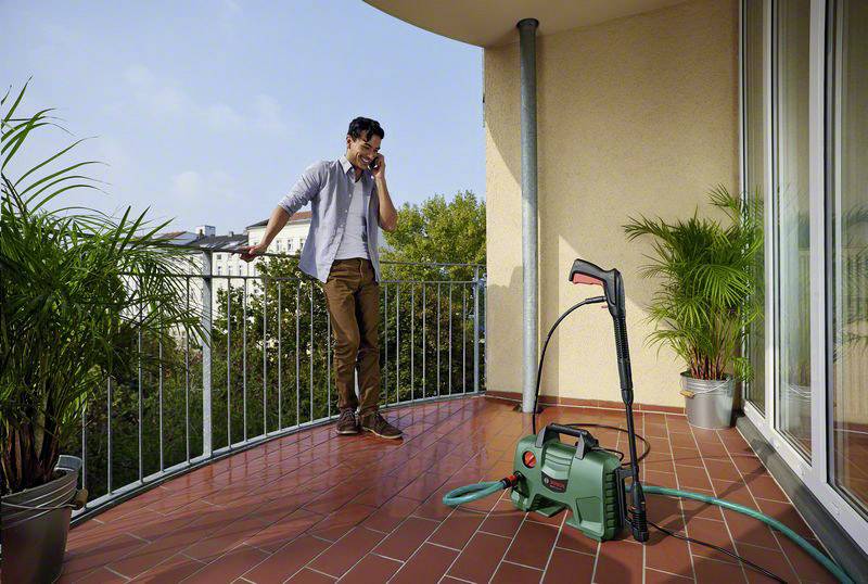 Un uomo sta telefonando su un balcone. Accanto a lui c'è un idropulitrice con tubo. Sullo sfondo sono visibili piante e edifici.