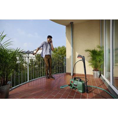 Un uomo sta telefonando su un balcone. Accanto a lui c'è un idropulitrice con tubo. Sullo sfondo sono visibili piante e edifici.