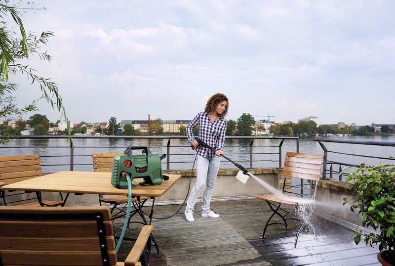 Una persona sta pulendo una terrazza con un idropulitrice. Sullo sfondo, un tavolo e sedie sono visibili accanto a una riva del fiume.
