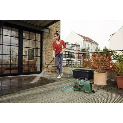 Donna pulisce una terrazza in legno con un idropulitrice. Vasi da fiori e ambiente urbano moderno sullo sfondo.