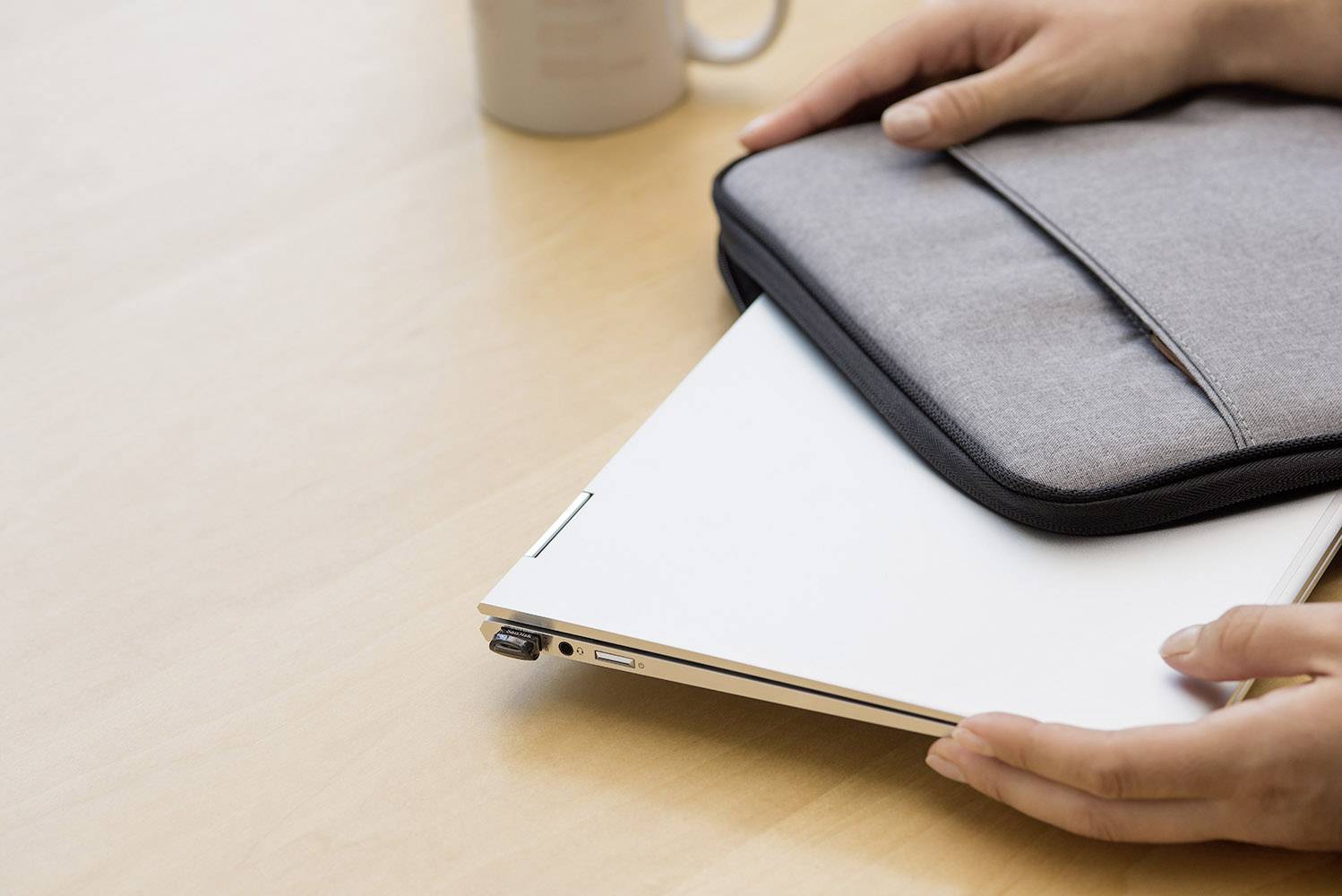 Una persona ripone un laptop chiuso in una custodia grigia su un tavolo di legno. Sullo sfondo, è presente una tazza.