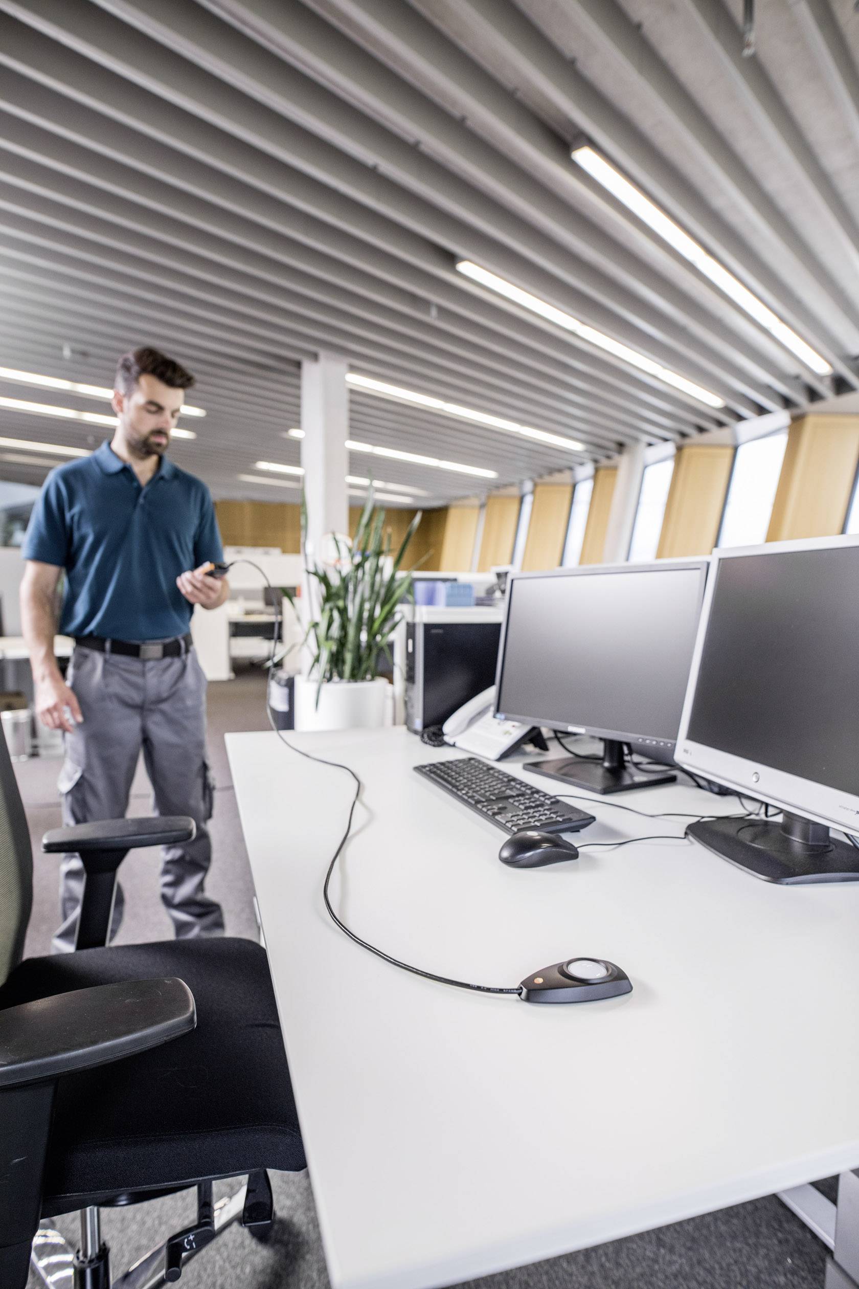 Un uomo in ufficio tiene in mano uno smartphone ed è in piedi accanto a una scrivania con due monitor, mouse e tastiera.